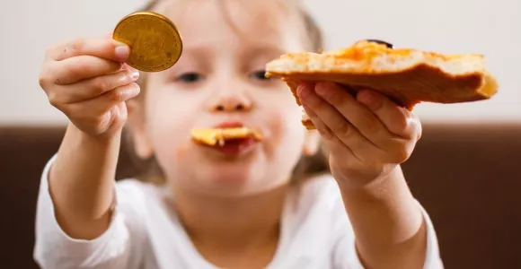 Enfant avec un bout de pizza dans la bouche, la part dans la main gauche, et un bitcoin dans la main droite.