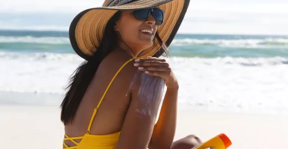 femme avec chapeau et lunettes de soleil, assise sur la place et s'enduisant de crème solaire.