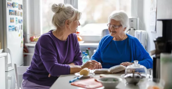 Dans une pièce de vie, une personne agée est attablée avec un livre et sourit à une personne plus jeune