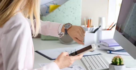 Une femme assise à un bureau regarde sa montre et tient un smartphone