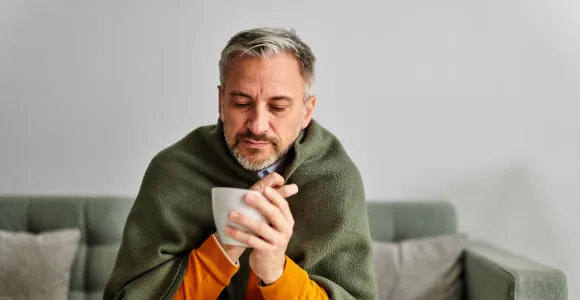 Un homme enrhumé est emmitouflé dans une couverture et a une tasse à la main.
