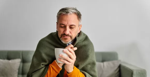 Un homme enrhumé est emmitouflé dans une couverture et a une tasse à la main.