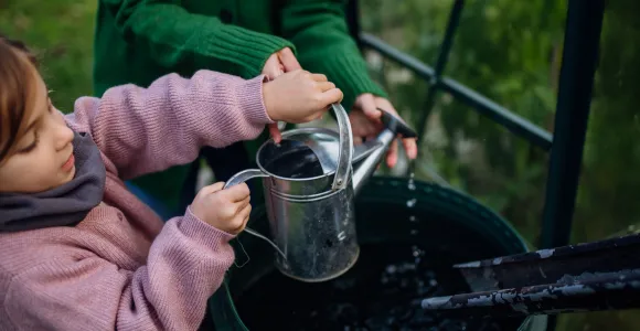 Une fillette et sa maman récupèrent de l'eau de pluie dans un arrosoir. 