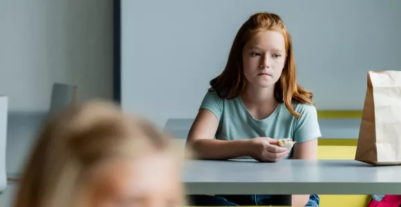 Une jeune fille mange une tartine toute seule au fond d'un réfectoire.