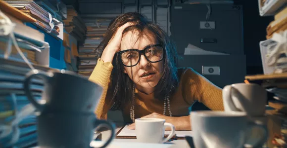 Une femme la tête dans une main accoudée sur un bureau, fatiguée et entourée de dossiers.