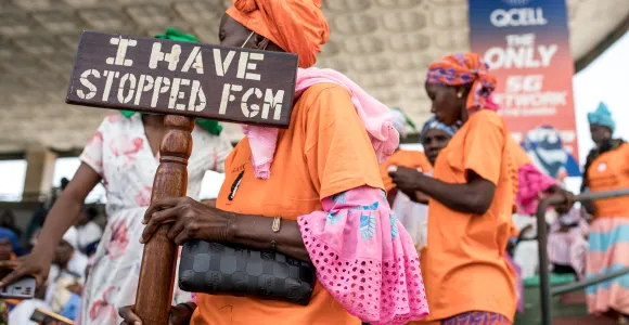 En mars 2024, une manifestante tient un panneau “Les MGF s’arrêtent avec moi” devant l’Assemblée Nationale de Gambie.