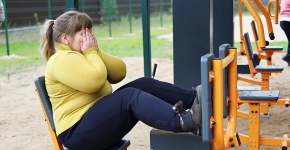Une ado en obésité assise sur une machine de sport à l'extérieur se tient le visage en pleurant