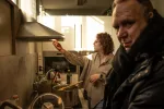 Une jeune femme en train d'allumer la hotte dans la cuisine du lieu. Un homme en avant-plan de la photo regarde vers l'appareil photo. 