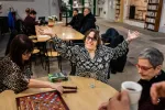Une bénéficiaire de la Croisée sourit, les bras ouverts. Elle joue au scrabble avec une autre bénéficiaire et une bénévole. 