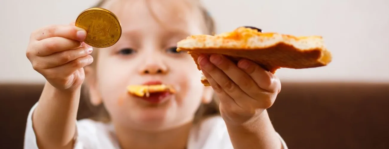 Enfant avec un bout de pizza dans la bouche, la part dans la main gauche, et un bitcoin dans la main droite.