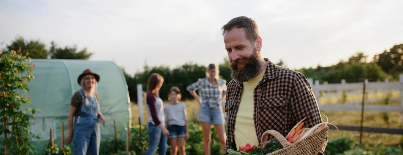 Personnes dans un champ cultivé dont l'homme en premier plan tient un panier en osier remplis de légumes.