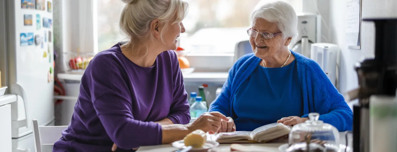 Dans une pièce de vie, une personne agée est attablée avec un livre et sourit à une personne plus jeune