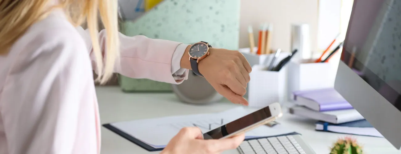 Une femme assise à un bureau regarde sa montre et tient un smartphone