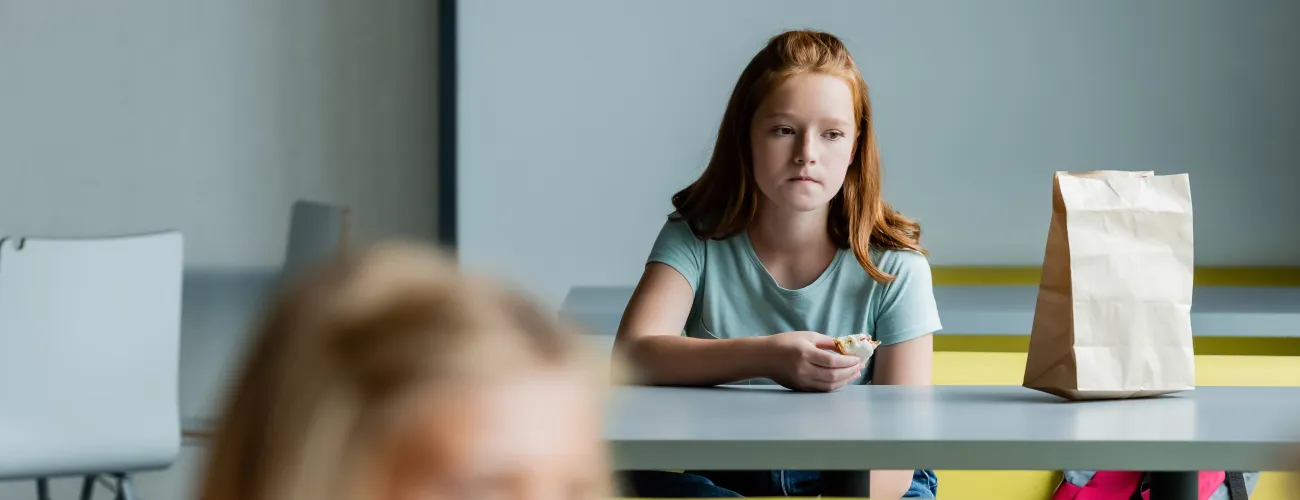 Une jeune fille mange une tartine toute seule au fond d'un réfectoire.