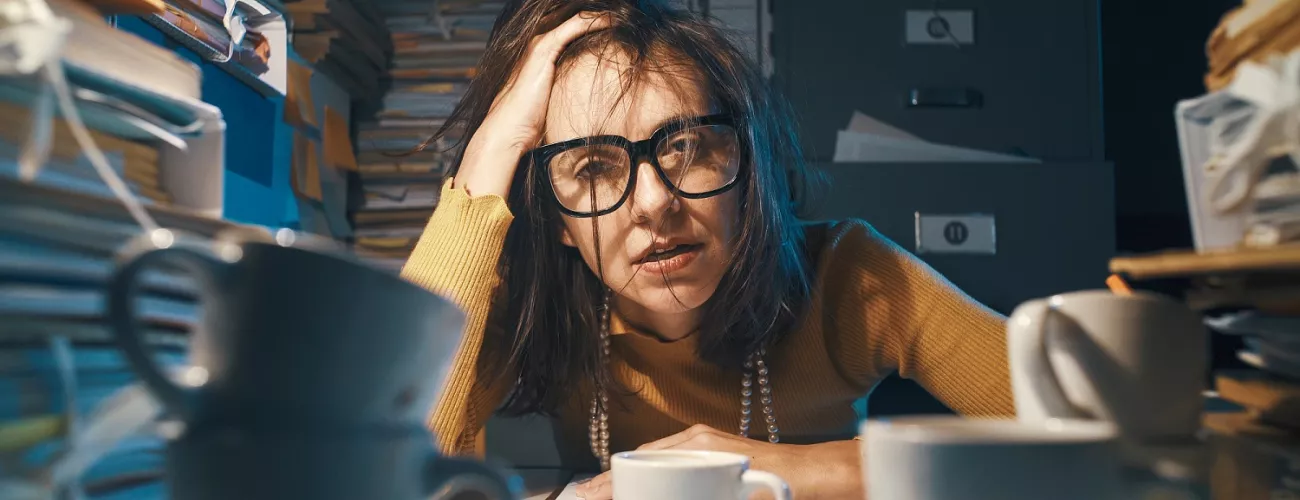 Une femme la tête dans une main accoudée sur un bureau, fatiguée et entourée de dossiers.