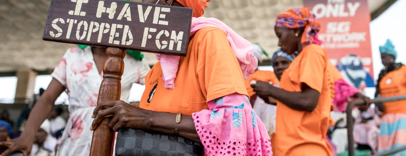 En mars 2024, une manifestante tient un panneau “Les MGF s’arrêtent avec moi” devant l’Assemblée Nationale de Gambie.