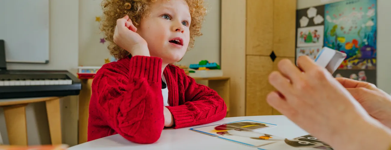 Enfant assis à une table, observant ce qu'un adulte lui montre.
