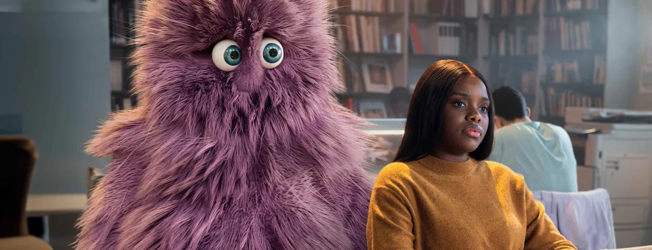 Une jeune femme assise dans un bibliothèque avec à l'arrière d'elle une grosse peluche
