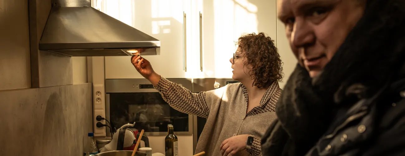 Une jeune femme en train d'allumer la hotte dans la cuisine du lieu. Un homme en avant-plan de la photo regarde vers l'appareil photo. 