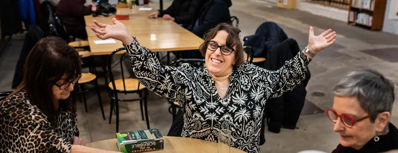 Une bénéficiaire de la Croisée sourit, les bras ouverts. Elle joue au scrabble avec une autre bénéficiaire et une bénévole. 