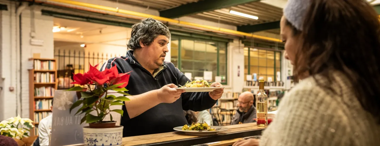 Héléna, bénéficiaire de la Croisée sert la salade liégeoise à une autre personne. 